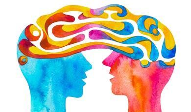 A vibrant watercolour painting showing two human profiles facing each other, their minds connected by a shared, swirling stream of bright yellow, pink, and blue patterns that form the shape of a single brain above them.