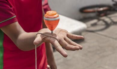 A close-up shot shows a person's open hand balancing a spinning orange and grey plastic top on their palm. The top is supported by a thick string wrapped around the fingers, while the person wears a red shirt with green trim