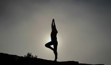 A silhouette of a person performing the yoga tree pose on a rocky hill against a hazy, backlit sky with the sun glowing behind them