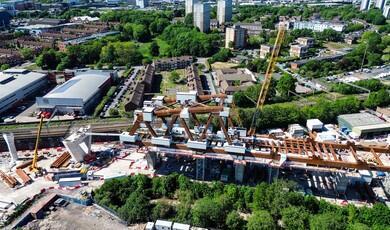 An aerial drone photograph capturing the extensive construction site of a massive steel truss bridge structure, surrounded by cranes, machinery, and urban residential buildings in the background