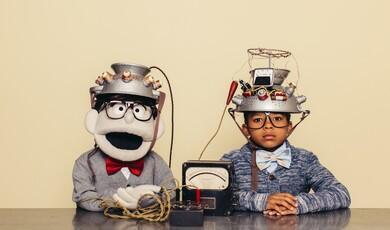 A young boy and a lookalike puppet sit side-by-side at a table wearing matching thick-rimmed glasses, bow ties, and elaborate, wire-covered colanders as "thought-reading" helmets connected to a vintage dial meter
