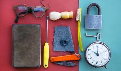 A flat-lay arrangement of vintage items, including a book, eyeglasses, a shaving brush, tools, a padlock with a key, and an alarm clock, neatly organised on a split red and teal background