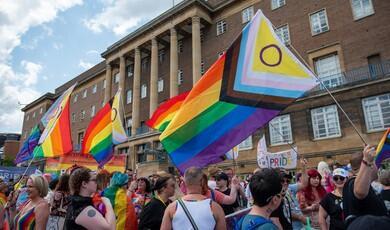 A wide shot captures a lively crowd during a Pride parade gathering outside a large, multi-story brick civic building featuring prominent classical columns