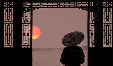 A person wearing a traditional Chinese conical hat stands before an ornate doorway, facing a calm lake at sunset.