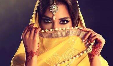 A close-up portrait of a South Asian woman looking directly at the camera, her face partially veiled by a vibrant yellow dupatta adorned with pearls, showcasing intricate henna designs on her hands and a maang tikka on her forehead