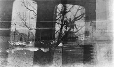 A high-contrast, black-and-white photograph taken through a window, creating a layered, double-exposure effect. Bare tree branches and the silhouette of a person riding a statue of a horse are framed within a window pane