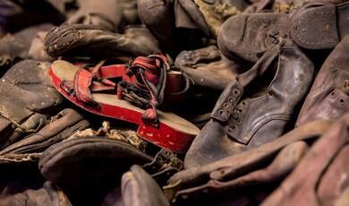 A large, tightly packed pile of old, worn, and dusty leather shoes and sandals belonging to victims of the Holocaust. Amidst the mostly dark brown and black footwear, a single pair of children's sandals with bright red wooden soles and frayed fabric straps stands out prominently near the centre
