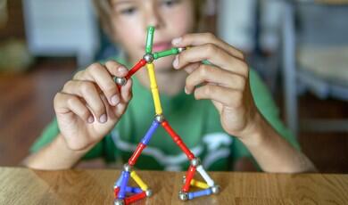 A young boy focuses intently on building a stick figure using a magnetic construction toy set made of colourful rods and metallic spheres on a wooden table