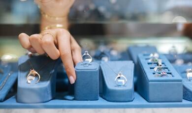 A close-up shot focuses on a person's hand with manicured nails pointing toward a solitaire diamond ring displayed on a blue velvet stand in a jewellery store showcase. The display features various fine jewellery pieces, including necklaces and rings, arranged neatly on matching blue velvet fixtures