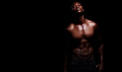 A shirtless, muscular man with dark skin poses against a black background, highlighted by dramatic low-key lighting
