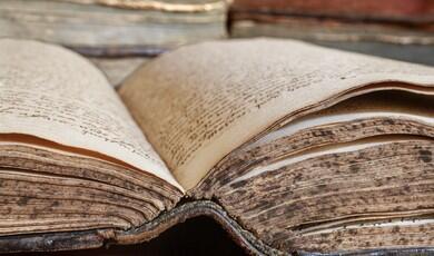 An open, ancient book with thick, weathered pages covered in handwritten script is shown in a close-up view