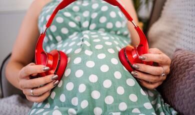 A close-up shot of a pregnant woman wearing a green and white polka dot dress, holding a pair of large, bright red over-ear headphones against her pregnant belly, positioning an ear cup on each side of the bump