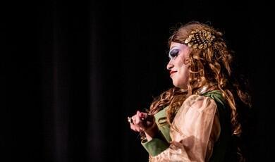 A profile shot of a performer with curly light brown hair, heavy stage makeup, and an ornate gold hair clip smiling while gesturing on a dark stage