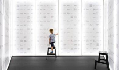 A young child stands on a black step stool with their back to the camera, reaching out to touch one of several large, floor-to-ceiling glowing white panels that form the walls of a room