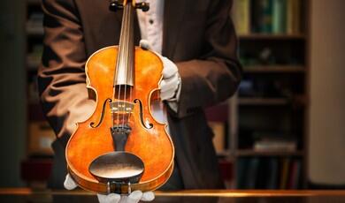 A close-up shot of a person in a brown suit jacket and white gloves holding out a polished wooden violin for display, against a blurred background of a wooden bookshelf filled with books