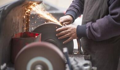 An artisan grinds a metal tool against a large, rotating bench grinder wheel, producing a bright shower of orange sparks. The worker, dressed in a dark apron and long sleeves, carefully guides the metal with bare hands. In the foreground, another grinding wheel sits out of focus on the workbench