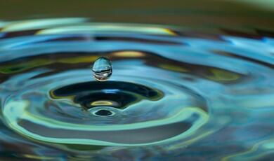 A close-up photograph captures a single water droplet suspended in the air just above a rippling water surface. The droplet reflects its colourful surroundings. Below it, concentric, circular ripples expand across the fluid surface, creating waves of light teal, deep blue, and pale yellow-green. The background shows a soft, out-of-focus blur of the same fluid colours