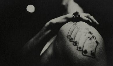 A high-contrast, grainy black and white photograph captures a person from a low angle, emphasising a tattoo on their upper arm and shoulder