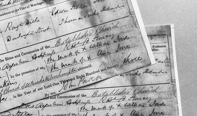 A close-up, black-and-white photograph of overlapping historical document pages, featuring elegant cursive handwriting on pre-printed forms