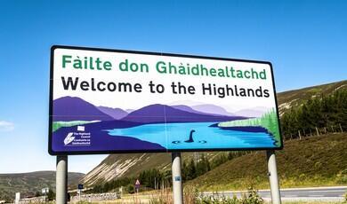 A large rectangular roadside welcome sign stands against a bright, clear blue sky, reading "Fàilte don Ghàidhealachd" in green text and "Welcome to the Highlands" in bold black text below it. The sign features an illustration of a scenic Scottish landscape with purple mountains, a blue lake, green pine trees, and a silhouette resembling the Loch Ness Monster