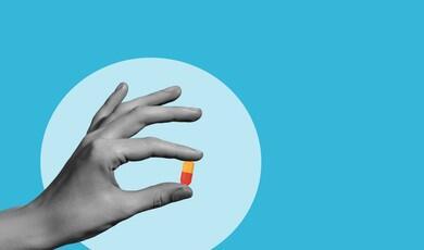 A stylised illustration features a black-and-white hand holding a small capsule pill against a bright blue background. The capsule is coloured with an orange top and a red bottom. A light blue circle serves as a backdrop behind the hand, highlighting the gesture
