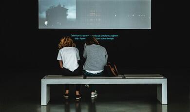 Two people sit on a bench from behind, watching a video projection with subtitles in a dark gallery space