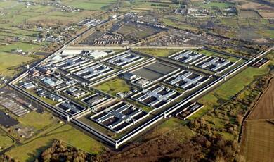 An aerial view of the HM Prison Maze complex, featuring its distinctive H-block layout with several H-shaped prison buildings surrounded by high security walls, watchtowers, and open green fields