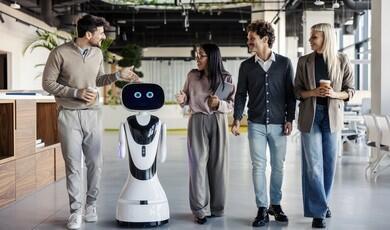 A small, futuristic white robot with digital blue eyes walks through a modern, open-plan office alongside four colleagues who are smiling and conversing