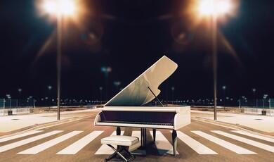 A white grand piano with its lid open sits in the middle of a pedestrian crosswalk on an empty city street at night, accompanied by a matching white piano bench. Two bright streetlights illuminate the scene from both sides, creating a dramatic, symmetrical glow against the dark sky