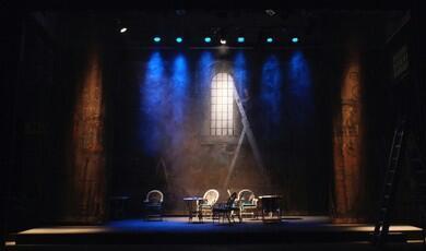 A theatre stage set designed to look like a stone wall courtyard or interior, illuminated by blue spotlights from above. In the centre backdrop, a painted arched window is lit with white light, with a ladder leaning against it. On the stage floor below, several wicker chairs and small round tables are arranged. Two large, dark columns frame the left and right sides of the stage