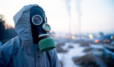 A person wearing a hooded protective jacket and a vintage gas mask stands outdoors against a blurred industrial background. The black rubber mask covers their entire face, featuring circular eye lenses, a perforated metal speech diaphragm, and a green cylindrical filter attached to the bottom. In the soft-focus distance, smoke plumes rise from industrial chimneys under a pale sky