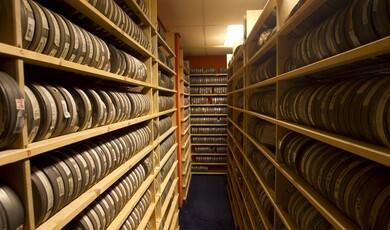 Two long rows of tall wooden shelves filled with rows of film canisters pack a narrow storage room with a blue carpeted floor. The shelves on the left and right extend forward into the foreground, while another shelf stands against the back wall