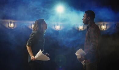 A young woman and man rehearse with scripts on a dimly lit theatre stage, illuminated by a bright spotlight amidst blue stage smoke