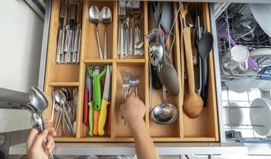 An overhead, eye-level shot captures a wooden kitchen drawer organiser filled with neatly arranged silverware and utensils, with a person's hands actively sorting forks and spoons