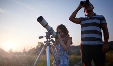 A father and his young daughter stargazing together in an open grassy field during sunset. The daughter looks through a white telescope mounted on a tripod, while her father stands beside her, looking up at the sky through a pair of binoculars