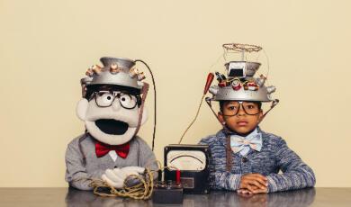 Two children, one real and one a puppet, wearing homemade metal helmets with wires sit at a table, connected to a small device.