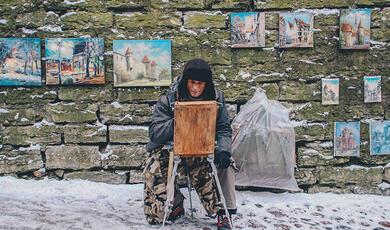 Man seated on street, painting, with other artworks on wall behind him