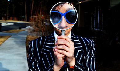 Man in pin stripe suit and sunglasses looking at the camera through a magnifying glass